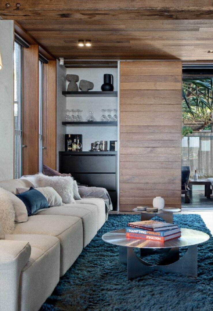 Timber-lined living room with a long sofa, a blue rug and shelves, opening to a garden courtyard