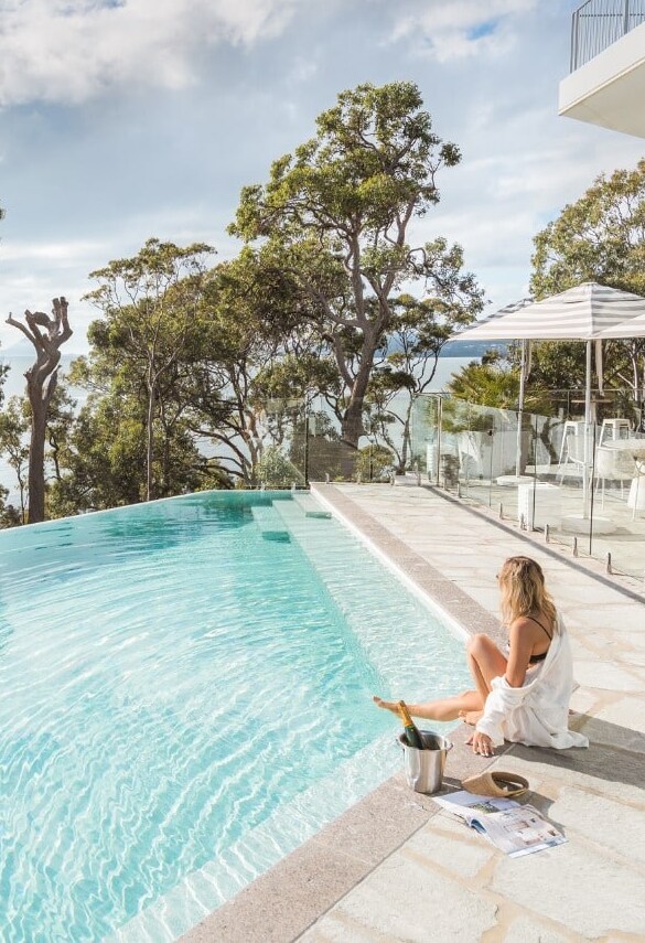 Woman sitting at the edge of an infinity pool beneath gum trees, a striped umbrella behind her