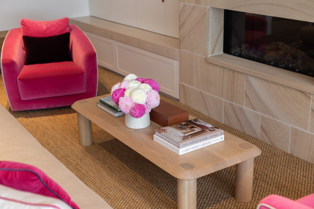 Oak coffee table with flowers and books between the pink armchair and the sandstone fireplace