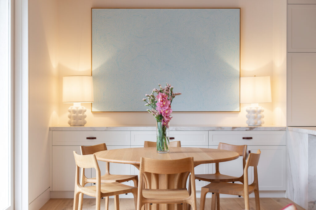 Round oak dining table beneath a large pale blue painting, with a lamp either side